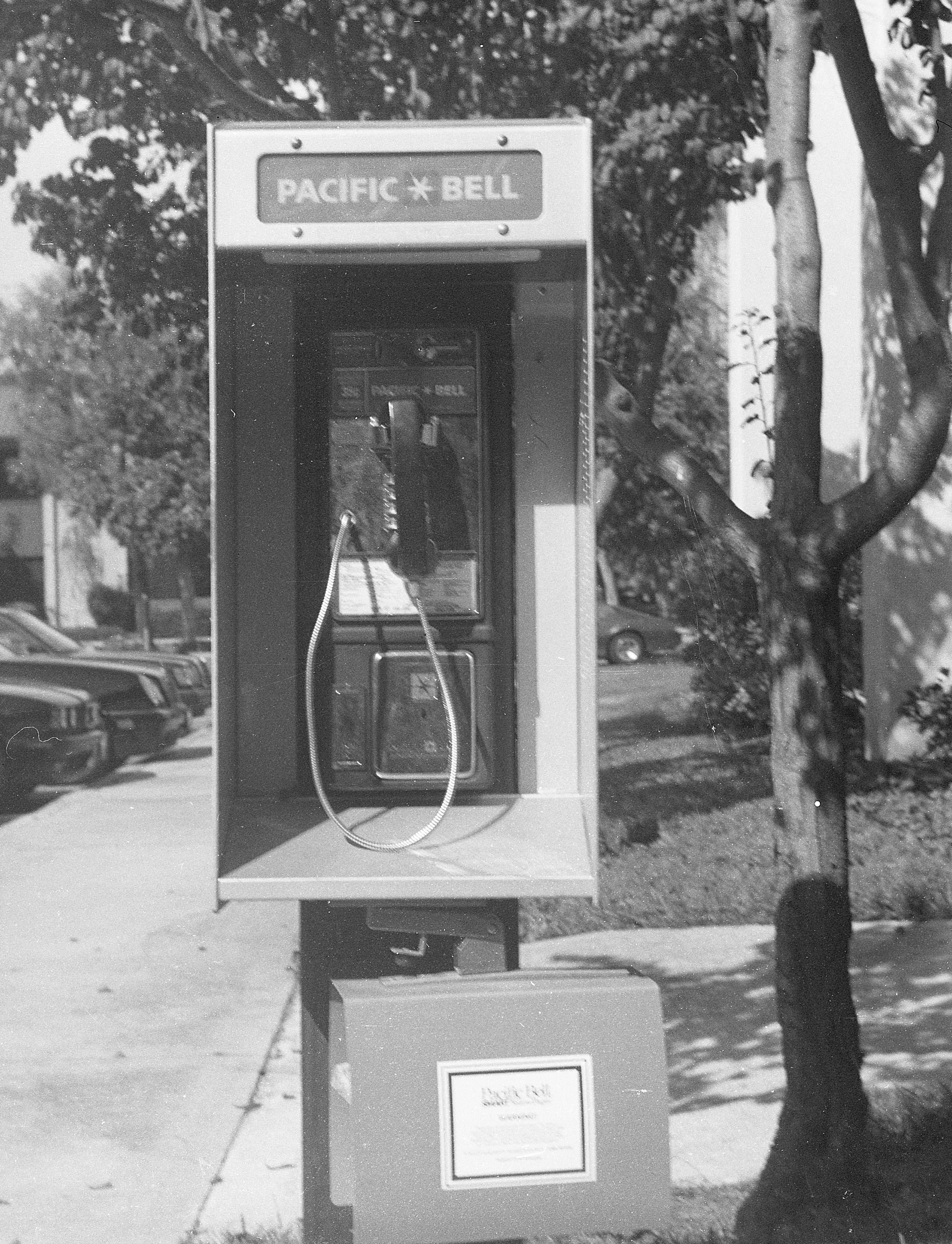 Black-and-white photograph of a Pacific Bell outdoor payphone in a booth on a sidewalk; parked cars line the curb and a tree frames the right edge.