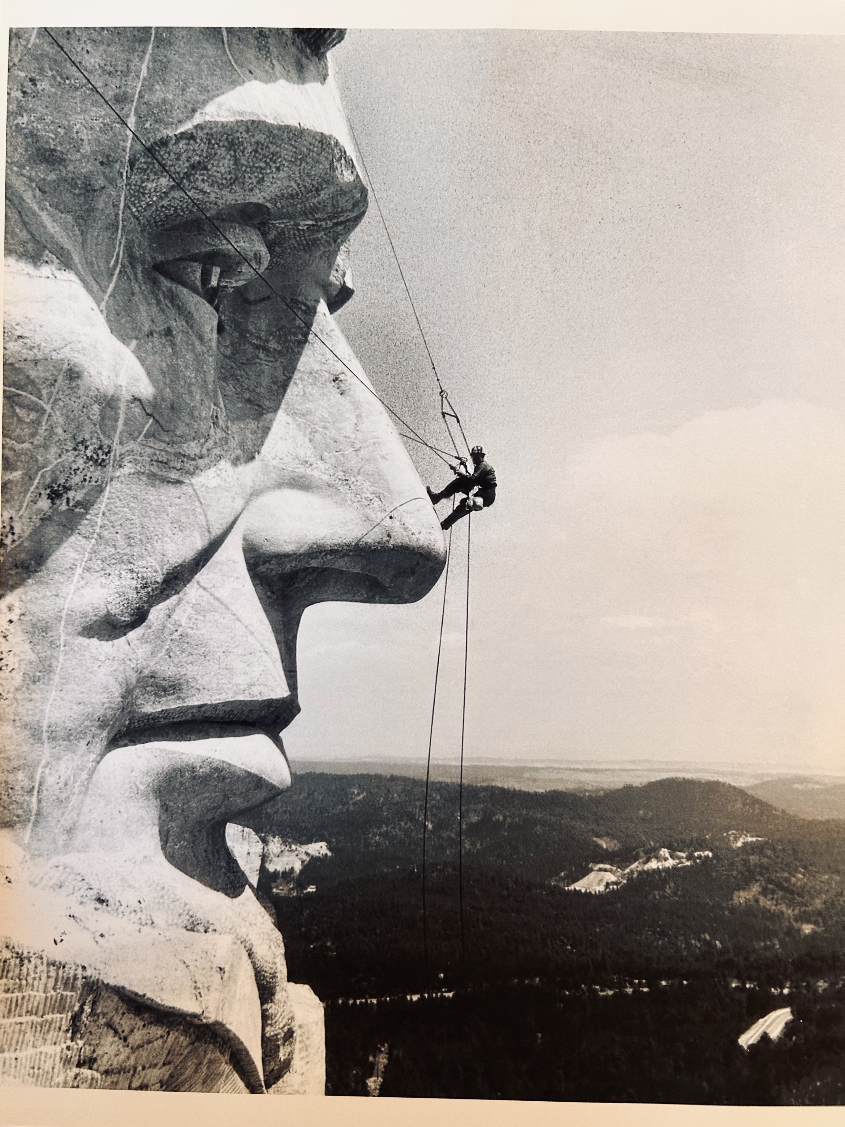 Black-and-white photograph of a craftsman suspended by ropes beside the face of Mount Rushmore, working near the nose of the carved stone figure with the Black Hills landscape below.